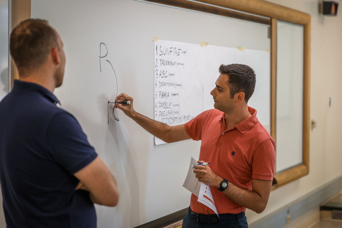 The image shows one man writing on a board while another observes.