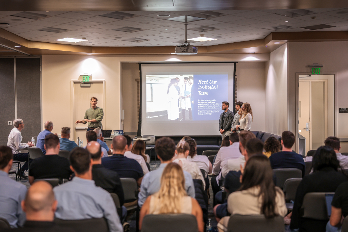 The image shows a team pitching their idea in front of a packed auditorium.