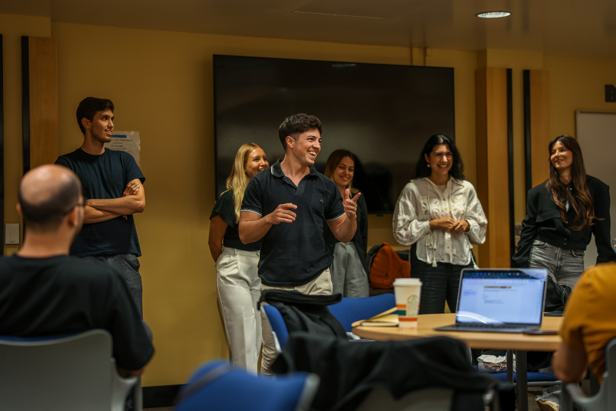 The image shows a group of people in front of a whiteboard presenting to others as part of a public speaking exercise.