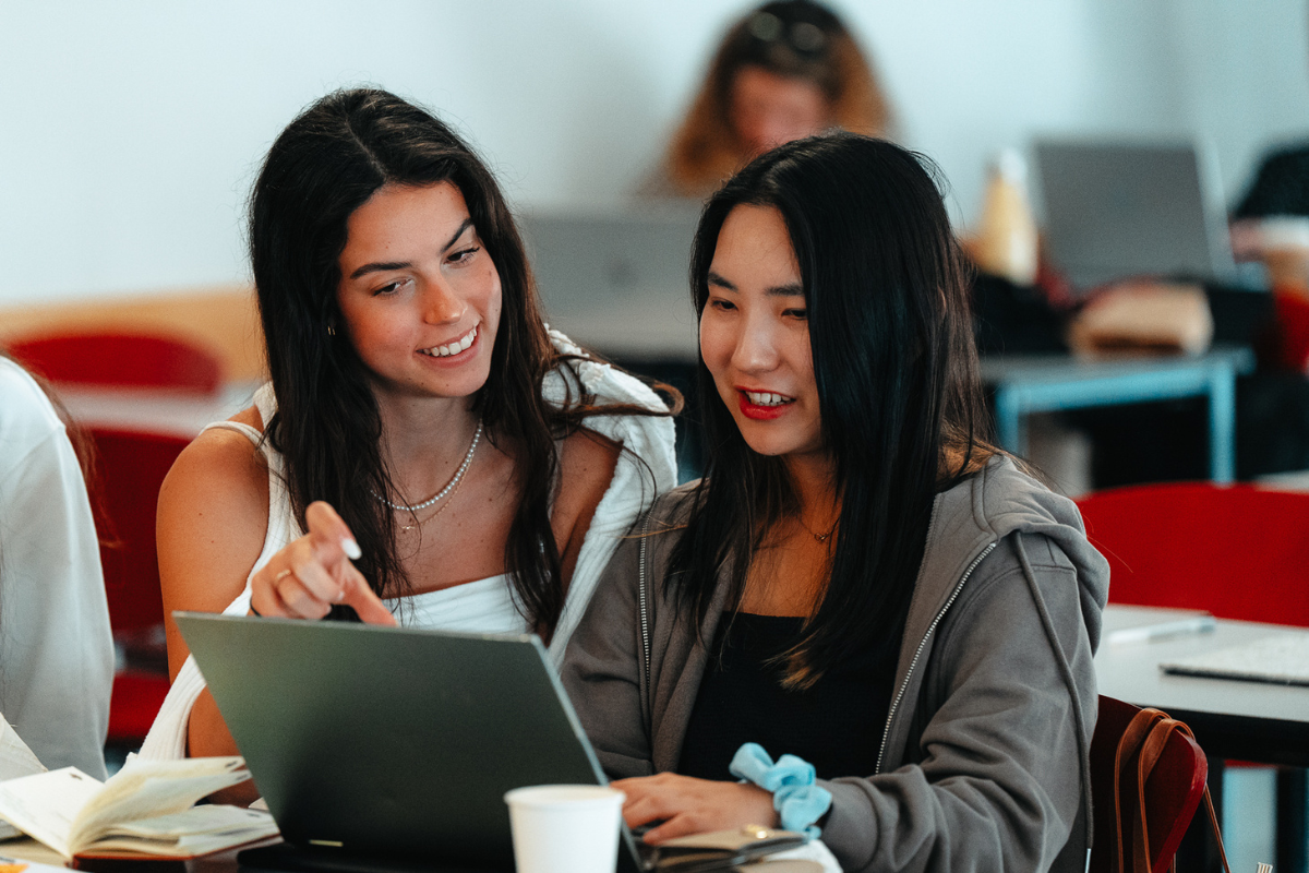 The image shows two women sitting in front of a laptop.